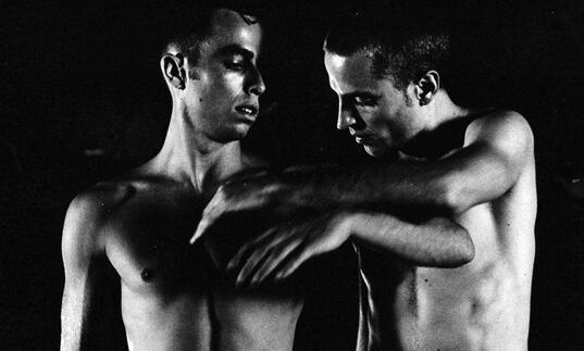 Two men stand topless in a dark room. 