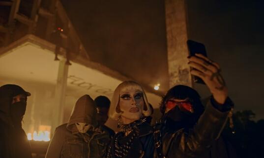 A woman with heavy makeup takes a selfie in front of a burning building, surrounded by masked people.