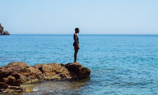 A woman in a bathing suit stands on a rock, looking out across the blue ocean in front of her.