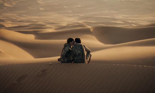 Two young people sit atop a sand dune in the vast expanse of the empty desert.