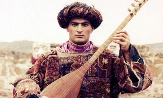 A young person playing a lute in traditional clothing, against a desert scape.