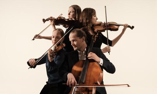 The Dudok Quartet playing their instruments. They are positioned in a close huddle, each person facing a different direction. They are against a cream coloured background.