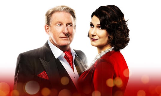 Adrian Dunbar and Stephanie J. Block pose alongside each other against a white and red background. Stephanie smiles down the camera, while Adrian looks at her cheekily. 