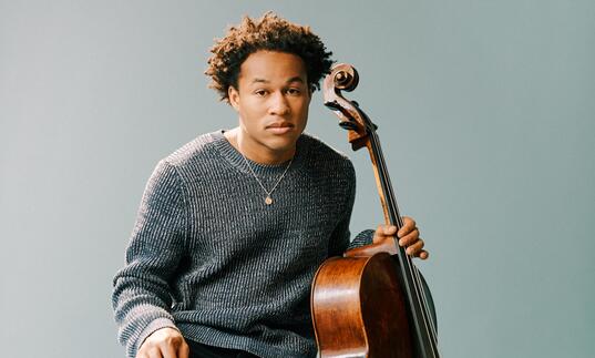 Sheku Kanneh-Mason sitting on a wooden stool with his cello, in front of a pale blueish gray background
