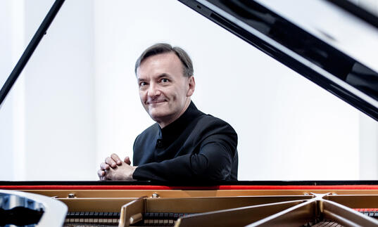 Stephen Hough sitting at a grand piano, smiling at the camera