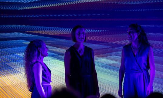 The three members of Voice Trio singing on stage, with purple lights and a purple projection behind them