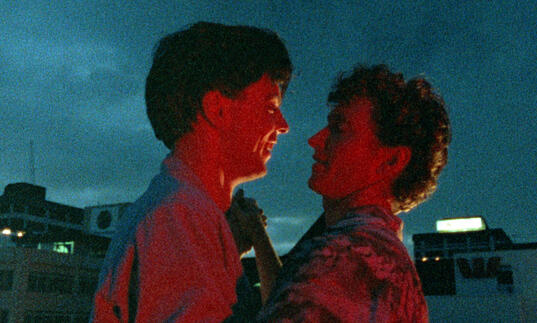 Two young men embrace each other in a dark city skyline. 