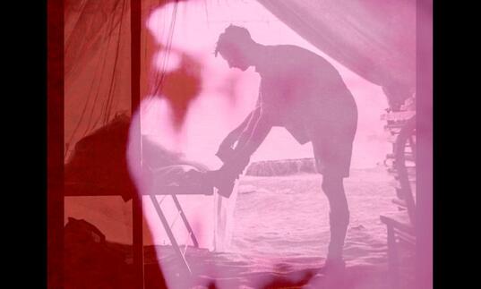 A pink and red image of a man tying his shoe lace in what looks like an army tent in a field. 