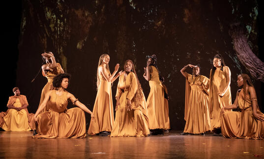 The cast of The Golden Stool performing on stage, wearing long golden dresses