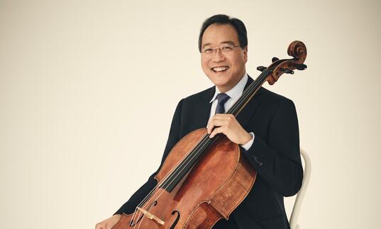 Yo-Yo Ma sitting with his cello, smiling at the camera, in front of a cream coloured background.