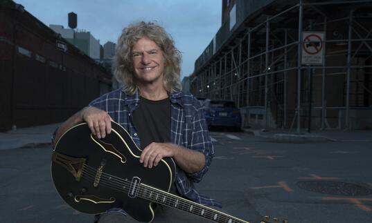 Pat Metheny sits, holding his guitar, on the street. 