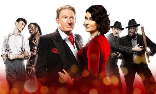 Adrian Dunbar and Stephanie J. Block stand side-by-side against a red background which has a yellow glow. The words 'Kiss Me, Kate, The Hit Musical Comedy' is written across it.