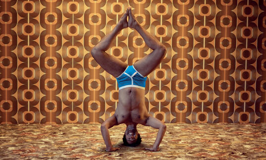 A person does a headstand in their pants against an orange and brown patterned wall.