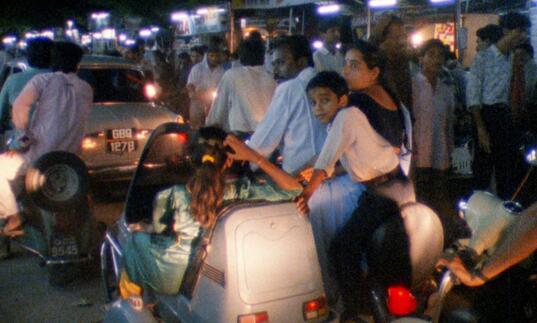 A family sit around a car on an urban street at night.
