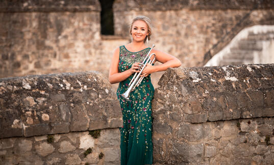 Matilda Lloyd in a green dress, holding her trumpet. She is stood within the walls of an old stone castle.