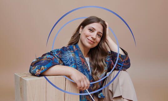 Dalia Stasevska sitting down, resting her arm on a wooden block. There are blue circular swirls around her.