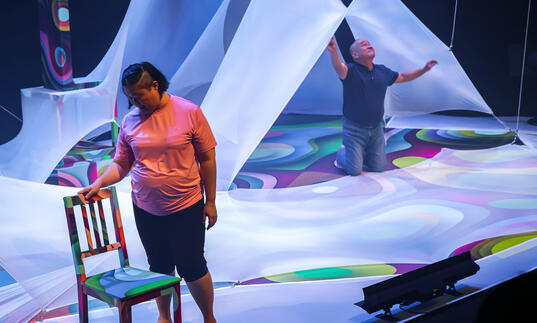 Two people perform on stage among large pieces of draping white fabric with bright lights projected onto it. 