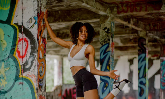 Writer Racheal Ofori poses, leaning against a concrete pillar while holding onto her heeled black shoe. 