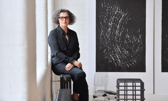 Mona Hatoum sitting on a radiator on the wall of her studio. On the wall next to her is Untitled (Bed Springs), one of her lithograph print depicting a structure made of bed springs, curved and crumpled.
