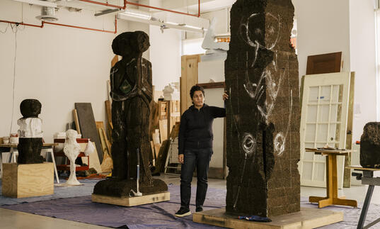 Huma Bhabha standing in a studio with two tall sculptures, one carved out and one with markings ready to be carved.
