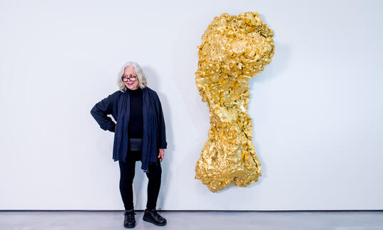 Lynda Benglis standing next to one her sculpture titled Ghost Dance/Pedmarks. It is a large, gold, torso-like form hanging on a white wall.
