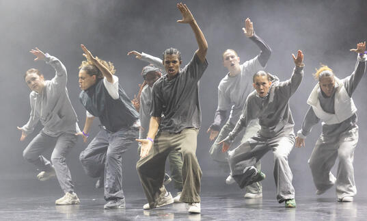 A group of hip hop dancers caught in motion. They lift one arm in the air and open their mouths.