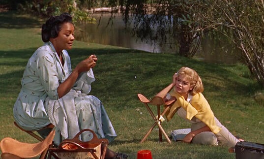 An older woman sits on the grass with a younger woman at her feet.