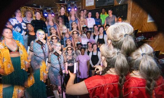 A choir group stands on stage in a dimly lit venue, with diverse singers wearing colorful outfits