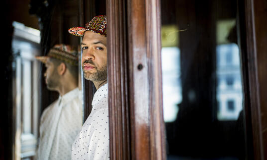 Jason Moran standing in a doorway. The back half of his body is obscured by the door frame, and he is turning to face the camera. His reflection is visible in a mirror behind him.
