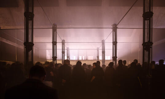 A large, smoke-filled room full of people, with two rows of large column speakers down the length of the room.