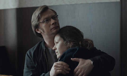 A man and woman hug in a grey room. 