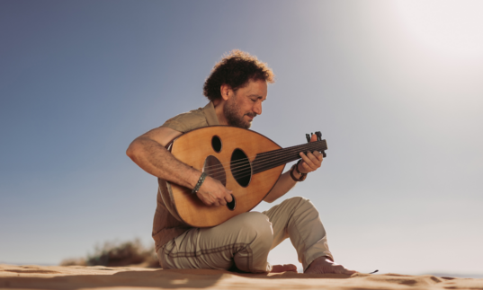 Portrait of Naseer Shamma sitting on sand and playing the oud