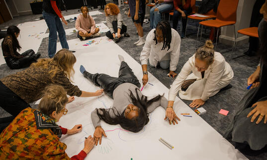 A person lies on the floor on a large piece of white paper. A group of people draw around them using a range of pens and pencils. 
