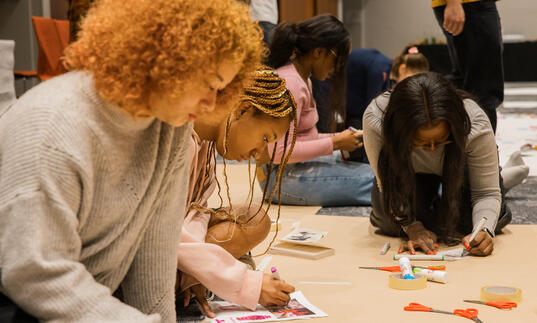 A group of people sit beside each other on the floor, drawing, colouring and collaging.