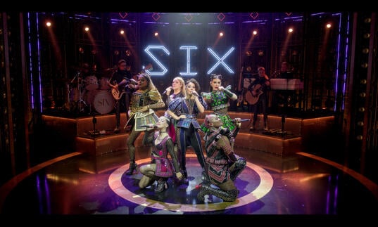 The queens perform on the stage with the band behind them. 