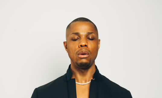 Davone Tines wearing a black suit with his eyes closed and hands in front of his chest
