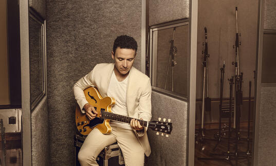 Image of Fonseca in a white suit sat down playing the guitar.