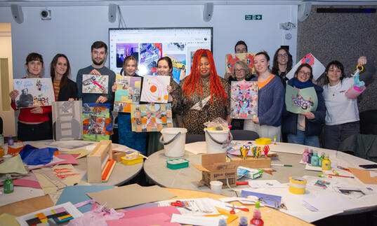 A group of people hold their artwork up proudly in front of a table filled with craft objects.