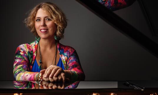 Gabriela Montero wearing a colourful dress sitting by a piano