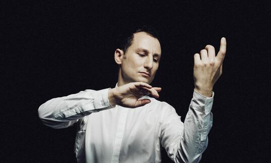 Violist Antoine Tamestit in front of a black background imitating playing his instrument without the actual instrument being there. His eyes are closed.