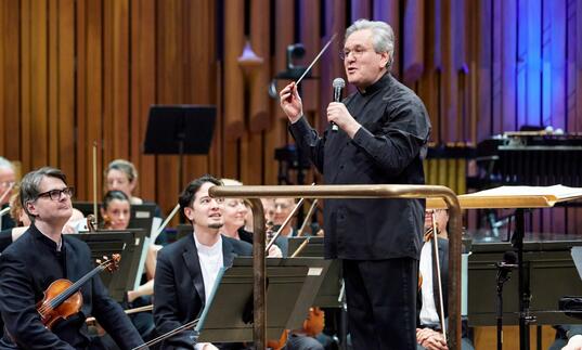 Sir Antonio Pappano standing on the podium facing the audience, talking to the audience, with the London Symphony Orchestra sat on-stage behind him