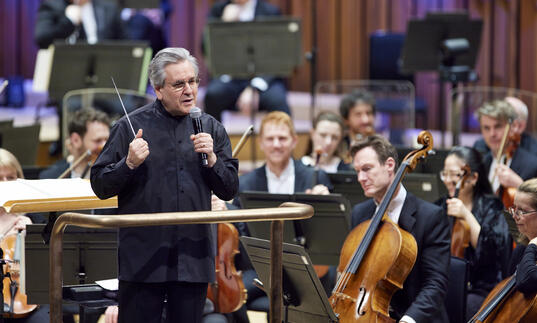 Sir Antonio Pappano standing on the podium on the Barbican stage, facing the audience and talking into a microphone