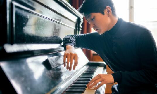Seong-Jin Cho sat at a piano, facing towards the keys, with his arm resting on the lid of the instrument
