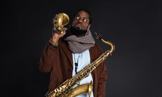 Soweto Kinch against a dark background, holding two saxophones
