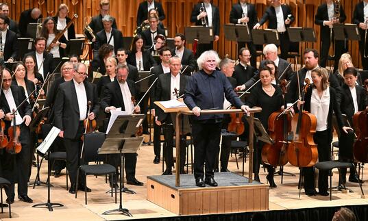 Sir Simon Rattle standing on the podium on the Barbican stage, facing the audience and smiling, with the London Symphony Orchestra standing behind him