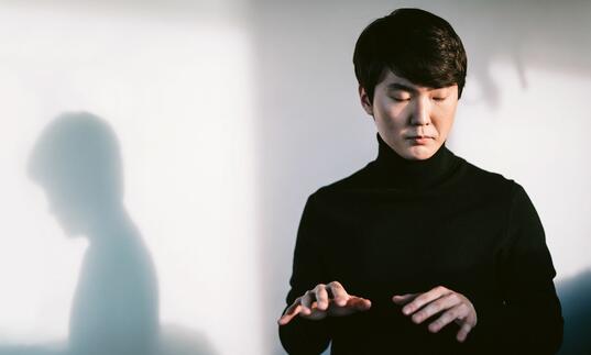Pianist Seong-Jin Cho in front of a white wall, with eyes closed lifting his hands as if playing the piano