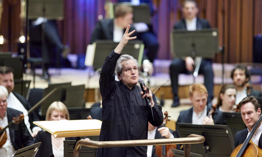 Sir Antonio Pappano standing on the conductor's podium, facing the audience, talking into a microphone with his right-hand raised