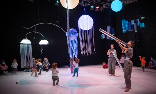 Young children run around a space interacting with large, colourful objects in a sensory show.