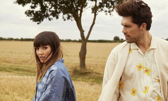 An image of both members of Oh Wonder walking away from each other in a field of grass with a tree behind them in the distance