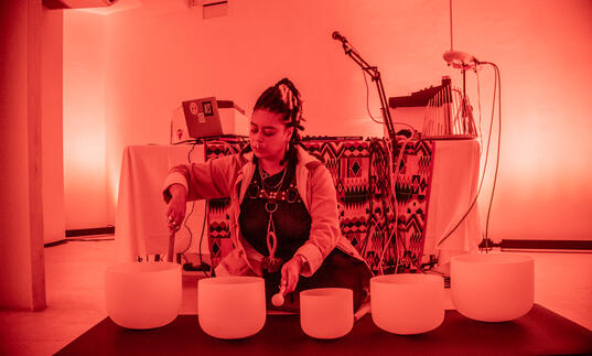 Hannah Catherine Jones sits in a room lit by an orange light playing bowl-shaped objects.